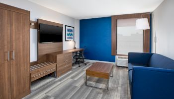 The image shows a modern hotel room with a blue accent wall, a sofa, desk, flat-screen TV, window with a lamp, and wooden furniture.
