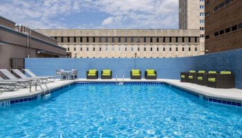 A rooftop swimming pool surrounded by lounge chairs and green-cushioned seating, with buildings in the background under a partly cloudy sky.